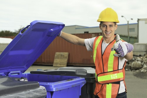 A young male waste collection worker wearing a yellow safety helmet, a high-visibility orange vest with reflective stripes, and purple gloves is standing outdoors in an industrial or commercial area. He is smiling and giving a thumbs-up gesture while holding the lid of a large blue plastic rubbish bin, which is partially open. Behind him, there are several other similar bins, some in shades of black and grey, arranged on a paved surface. In the background, there are warehouses, a red wooden fence, and a few scattered pallets, indicating a setting related to waste management or rubbish removal services. The environment appears overcast with natural daylight illuminating the scene, emphasizing the professional approach of Business Waste Removal St Johns Wood in providing waste disposal solutions that may serve local businesses and properties in the St Johns Wood area near London.