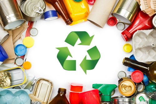 A collection of various recycled waste items arranged in a circular pattern around a central green recycling symbol. The waste includes metal cans with ribbed and smooth finishes in silver, red, and yellow tones, plastic bottles in green, yellow, and amber colors, paper and cardboard tubes with textured surfaces, and crumpled white paper or foam. The items are scattered on a plain white background, creating a contrast that highlights the textures and colours. The scene appears to be set outdoors or on a neutral surface, emphasizing environmental sustainability and waste management themes relevant to rubbish removal services in St Johns Wood. Business Waste Removal St Johns Wood uses this visual to represent their focus on recycling and responsible disposal of rubbish, reflecting both local environmental commitments and the broad category of waste types handled in their services.