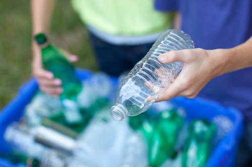 A close-up view of a person's hand holding a clear plastic water bottle with the cap removed, positioned above a blue recycling bin filled with various discarded bottles, containers, and plastic waste. In the background, another individual in a high-visibility vest is partially visible, with their hand resting on the edge of the bin, suggesting a rubbish collection or recycling activity. The scene appears to take place outdoors on a grassy area, possibly near a residential or commercial property in St Johns Wood, with natural daylight illuminating the scene. The plastic waste includes green and transparent bottles, some crushed or partially flattened, indicating the sort of materials handled during rubbish removal services provided by Business Waste Removal St Johns Wood. The focus is on the plastic waste, emphasizing environmentally conscious disposal and recycling efforts in the local area.