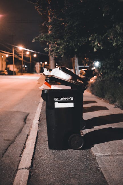 A collection of mixed waste materials dumped at the base of a brick and concrete wall; the pile includes flattened cardboard boxes, some partially crushed and open, alongside plastic bags filled with 